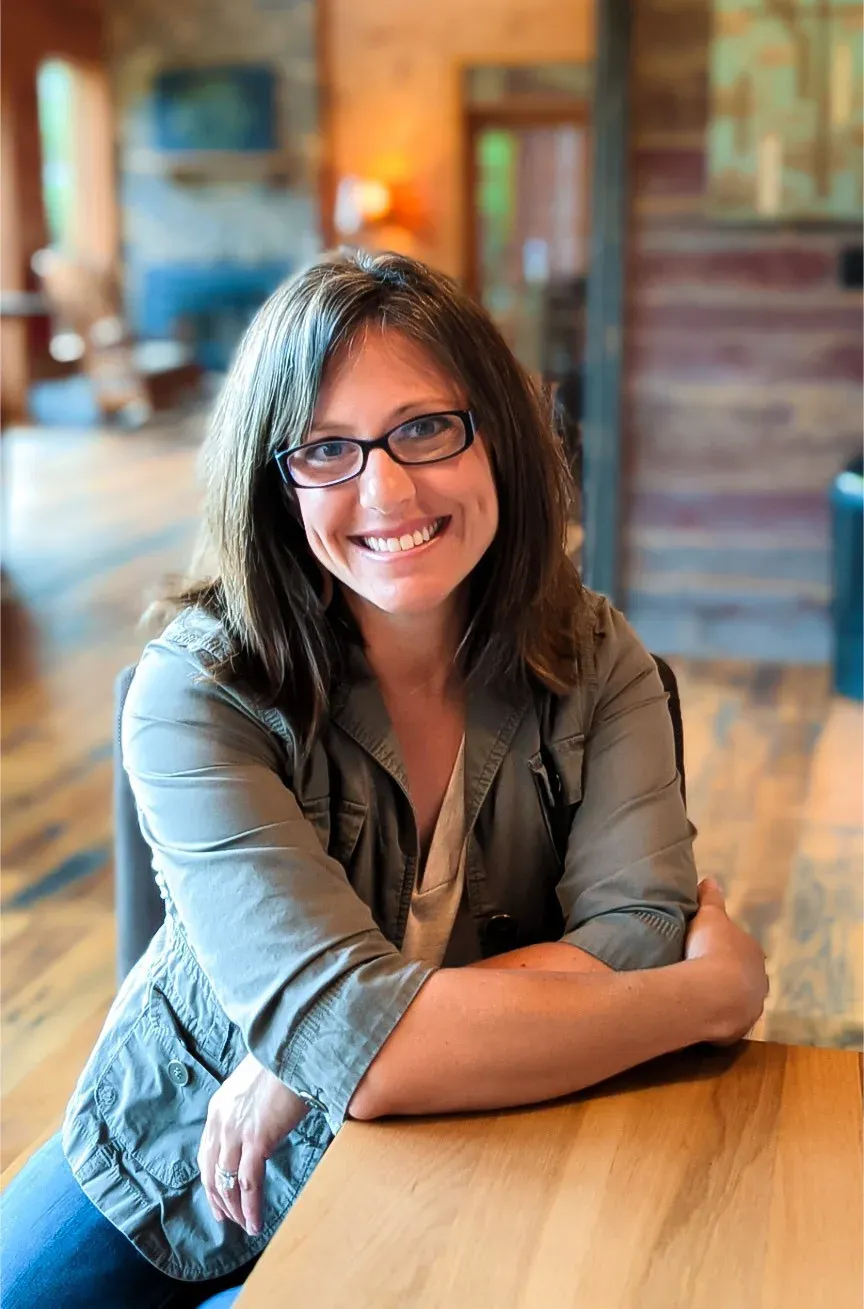 Kasie Secrest smiling, seated at a wooden table in a sunlit room, wearing glasses and an olive jacket