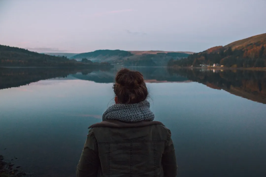 A woman standing calmly by a lake at dawn, looking out over the water